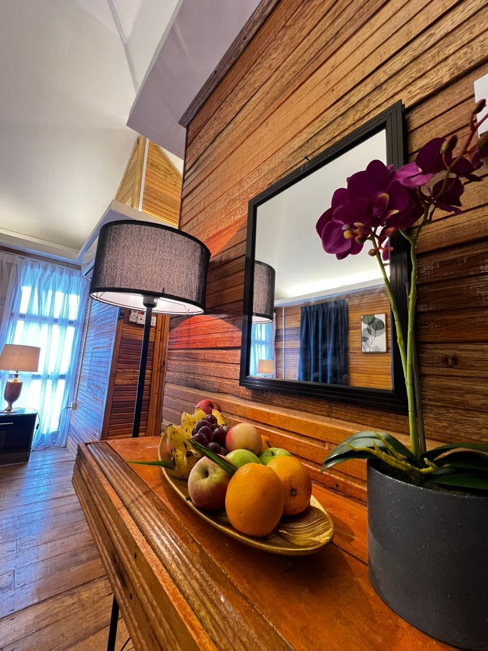 A wooden-framed room with a mirror, a lamp, a fruit bowl, and a potted plant.