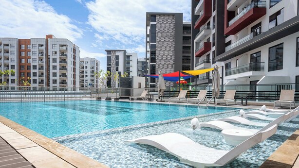 Une piscine avec des chaises longues, un parasol coloré et des bâtiments modernes en arrière-plan.