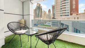 A balcony with a glass railing, two wicker chairs, a small table, and a view of a cityscape with tall buildings. - StayShort - Chic 1BR Sleeps 2 in Dubai Marina Near Metro (Dubai Marina)