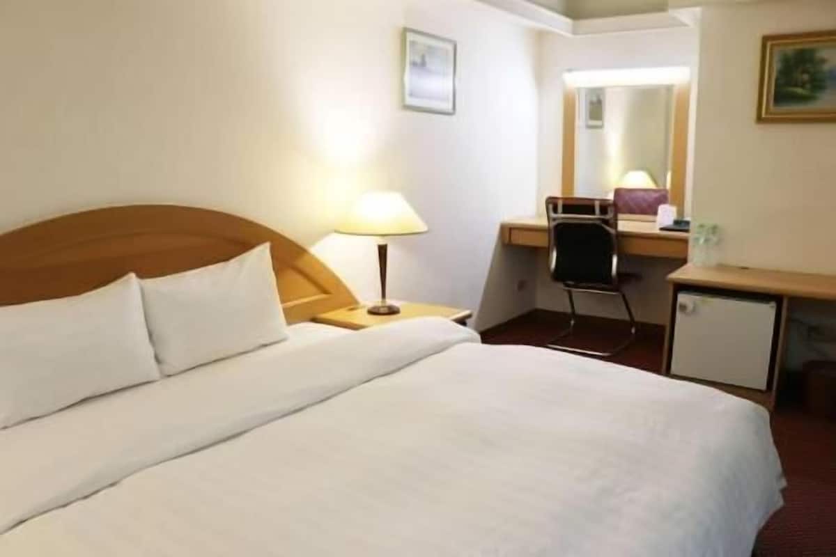 Una habitación de hotel con cama, escritorio, silla, lámpara y un mueble pequeño.