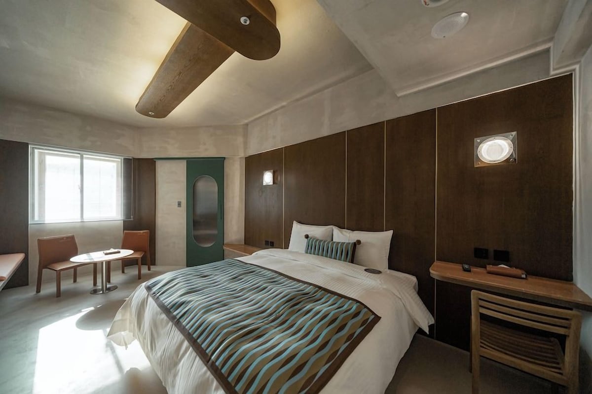 A modern hotel room with a large bed, a small round table, a seating area with chairs, and a large window providing natural light.