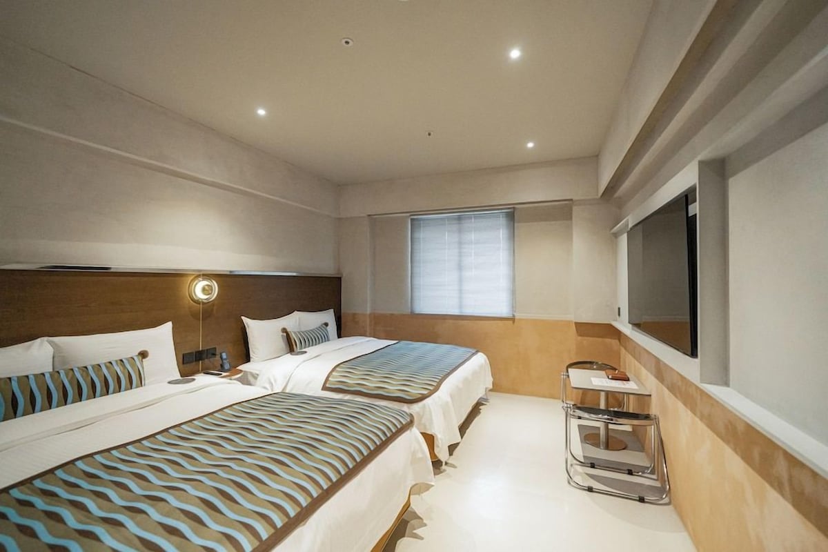 A hotel room with two beds, a bedside table, a lamp, a TV, and a window.