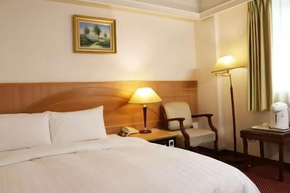 Habitación de hotel con cama, una silla, un escritorio con lámpara, teléfono y un cuadro en la pared.