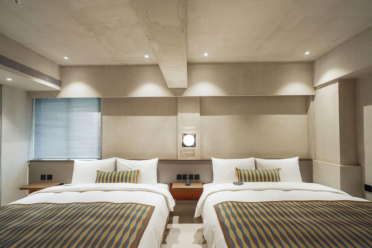 A hotel room with two beds, a nightstand, a wall-mounted lamp, and a window with blinds.