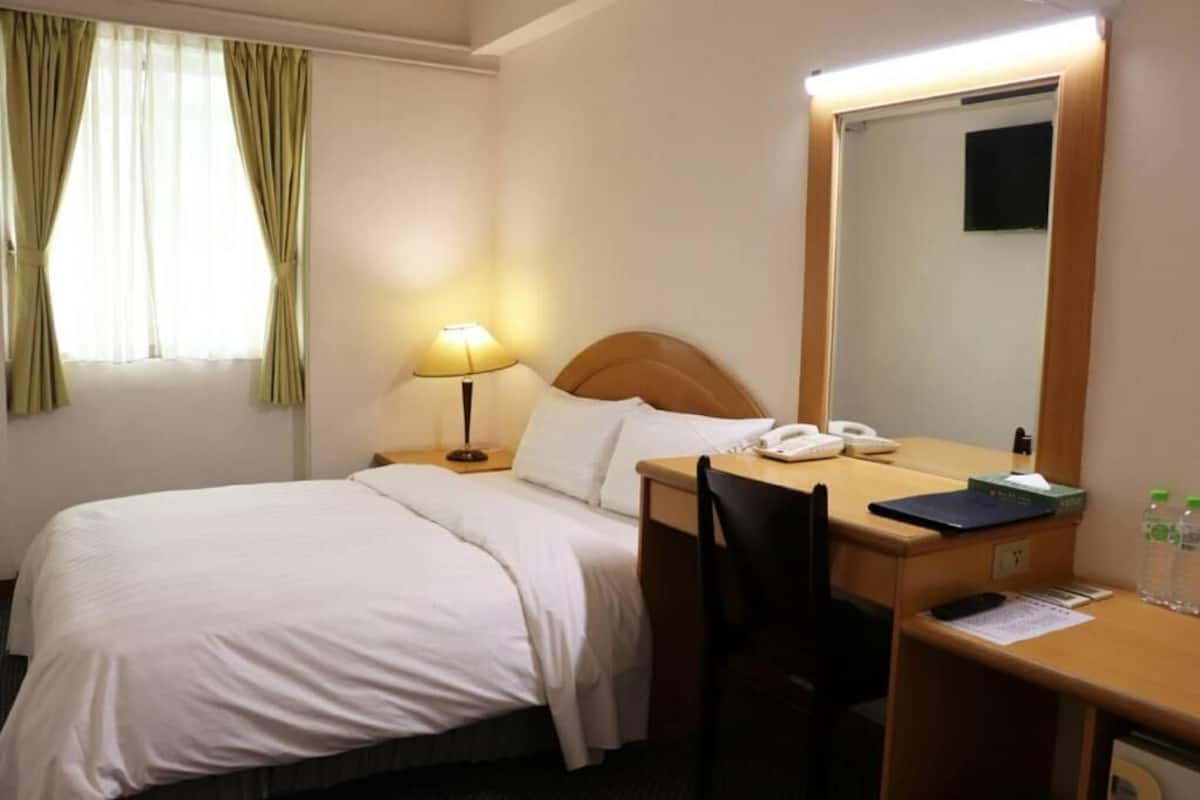 Habitación de hotel con cama, escritorio con lámpara, silla, espejo, teléfono y pequeña mesa con cuaderno y botellas de agua.