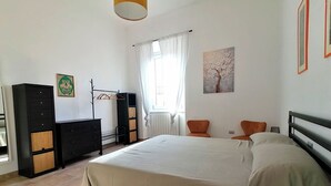 Une chambre à coucher comprenant un lit, une armoire, un porte-manteau, deux chaises, une fenêtre avec des rideaux et un tableau accroché au mur.