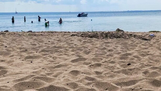Ein sandiger Strand mit Menschen im Wasser, Booten in der Ferne und einem klaren Himmel.