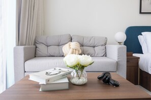 A modern living room with a grey sofa, a glass vase with white flowers, a book, and a remote control on a wooden coffee table. - Bloomfields Studio in The Gate - Masdar (Abu Dhabi)