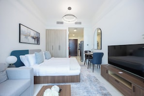 A modern hotel room with a bed, a sofa, a TV, a dining area, and a bathroom visible through an open door. - Bloomfields Studio in The Gate - Masdar (Abu Dhabi)