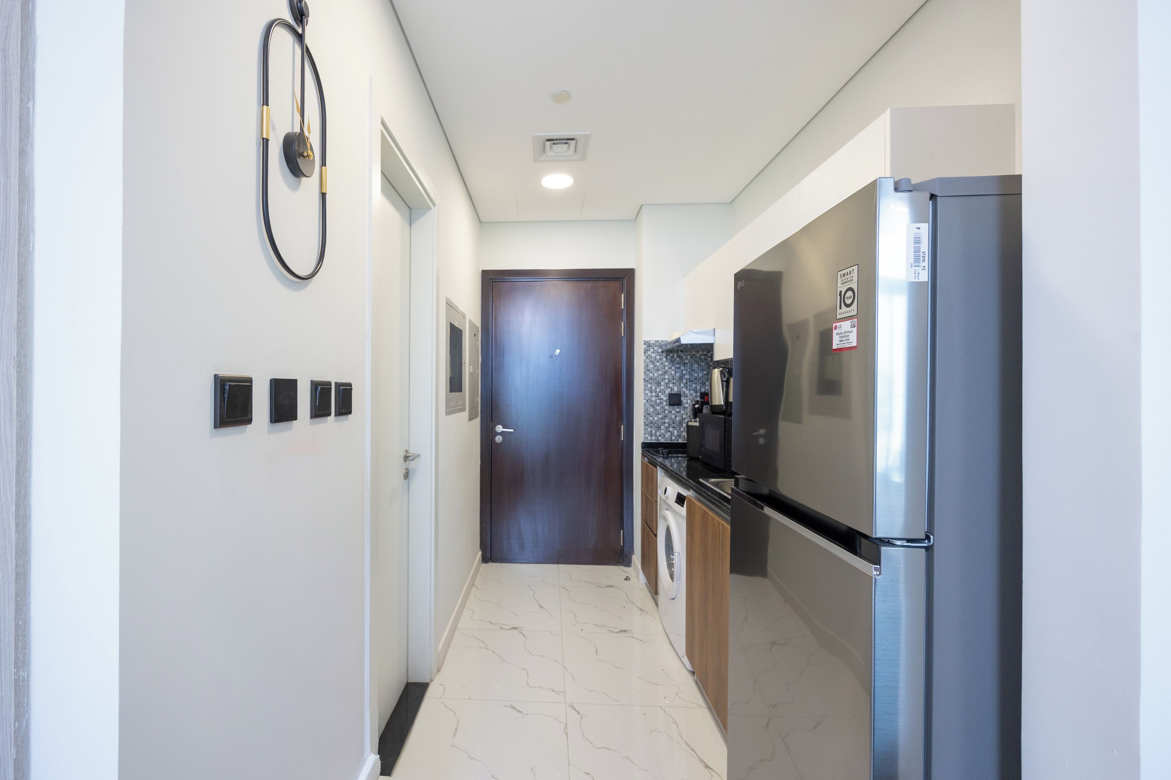 A narrow hallway with a door at the end, a wall-mounted clock, and a refrigerator on the right.