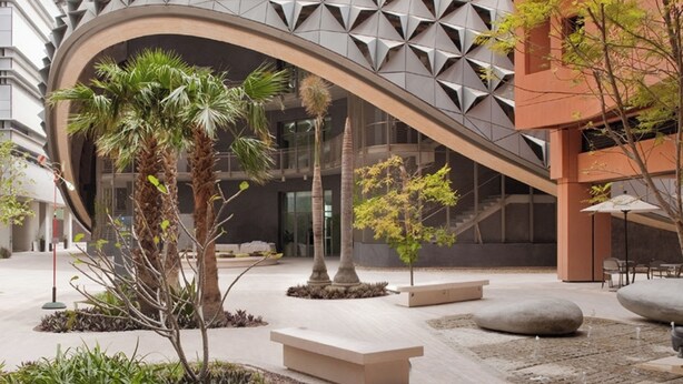 A modern building with a unique geometric facade, surrounded by a landscaped courtyard with palm trees and seating areas. - Bloomfields Vibrant Stu in The Gate (Abu Dhabi)