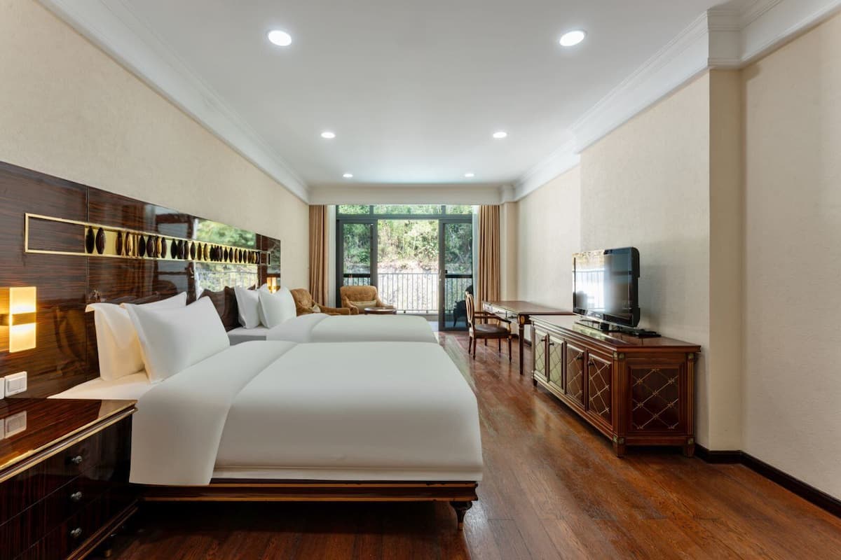 Habitación de hotel con dos camas, televisor, mueble de madera y balcón con vistas a zonas verdes.