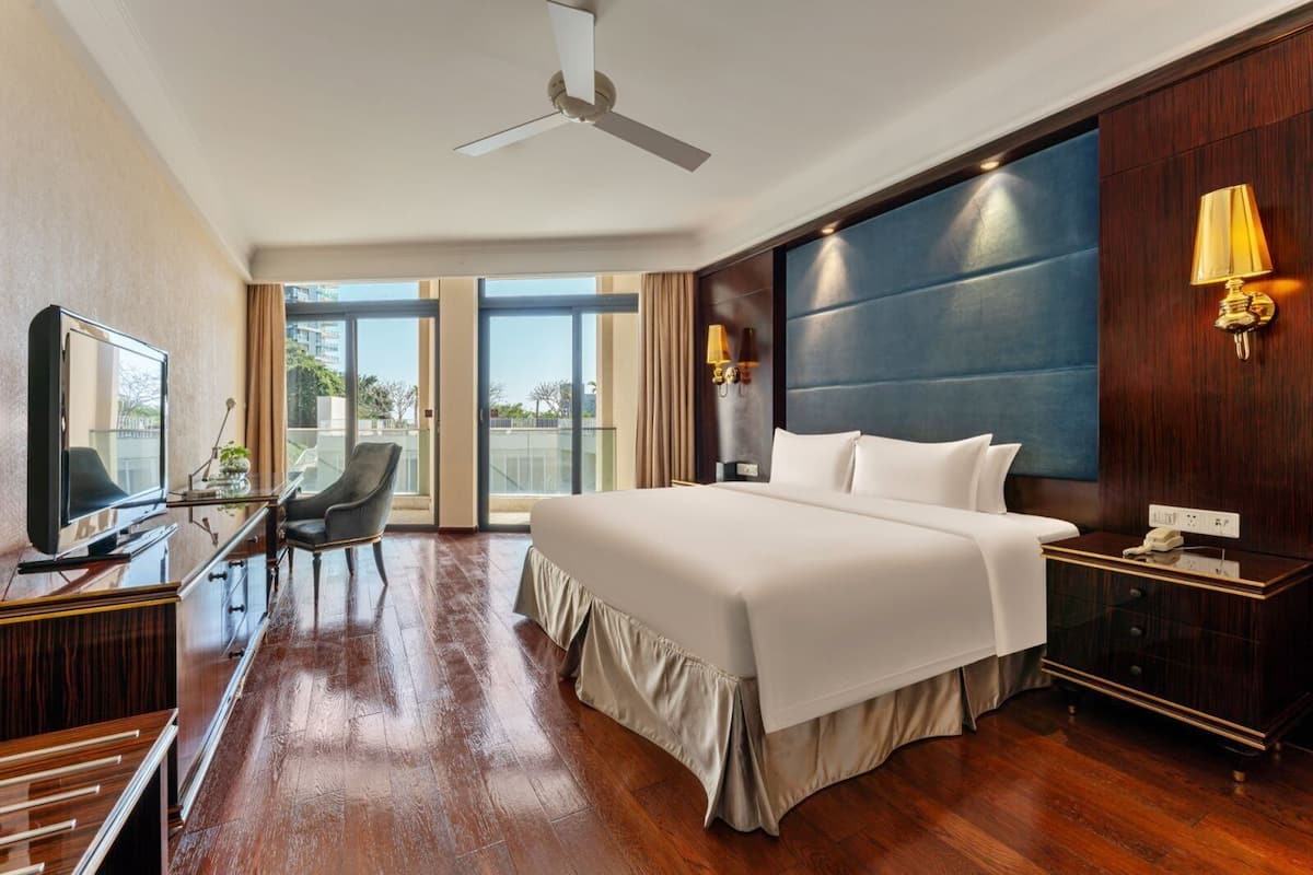 Habitación de hotel con cama, escritorio con silla, televisor, ventilador de techo y un ventanal grande.