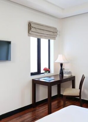 Habitación de hotel con cama, escritorio con lámpara, silla, ventana con cortinas y televisor de pantalla plana montado en la pared.