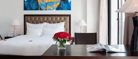 A hotel room with a bed, a bedside table with a lamp, a vase with flowers, and a painting on the wall.