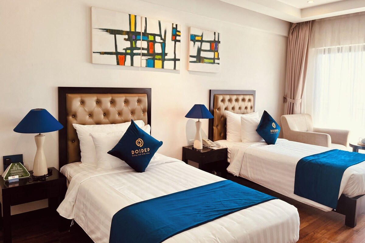 Habitació d'hotel amb dos llits, cadascun amb un llençol blau i almoines a joc. Hi ha dues taules de nit amb làmpades, una cadira i una obra d'art a la paret.