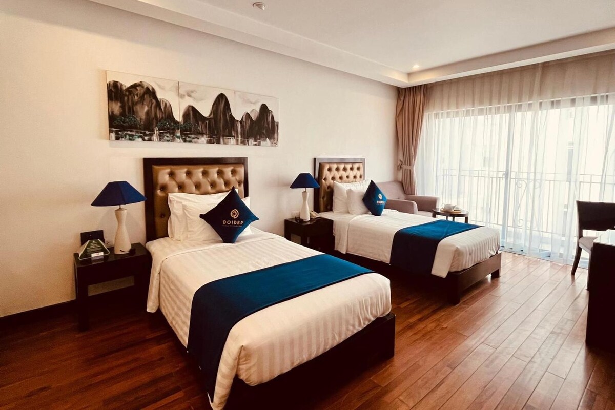 Habitació d'hotel amb dos llits, un finestral gran amb cortines, un quadre a la paret i un terra de fusta.