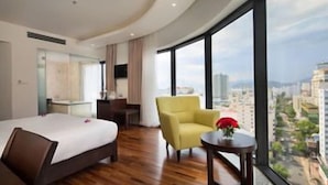 Een hotelkamer met een bed, een stoel, een klein tafeltje met een vaas en een groot raam met uitzicht op de stad.