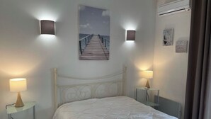 A neatly arranged bedroom with a bed, two bedside tables with lamps, a wall-mounted picture, and an air conditioning unit.