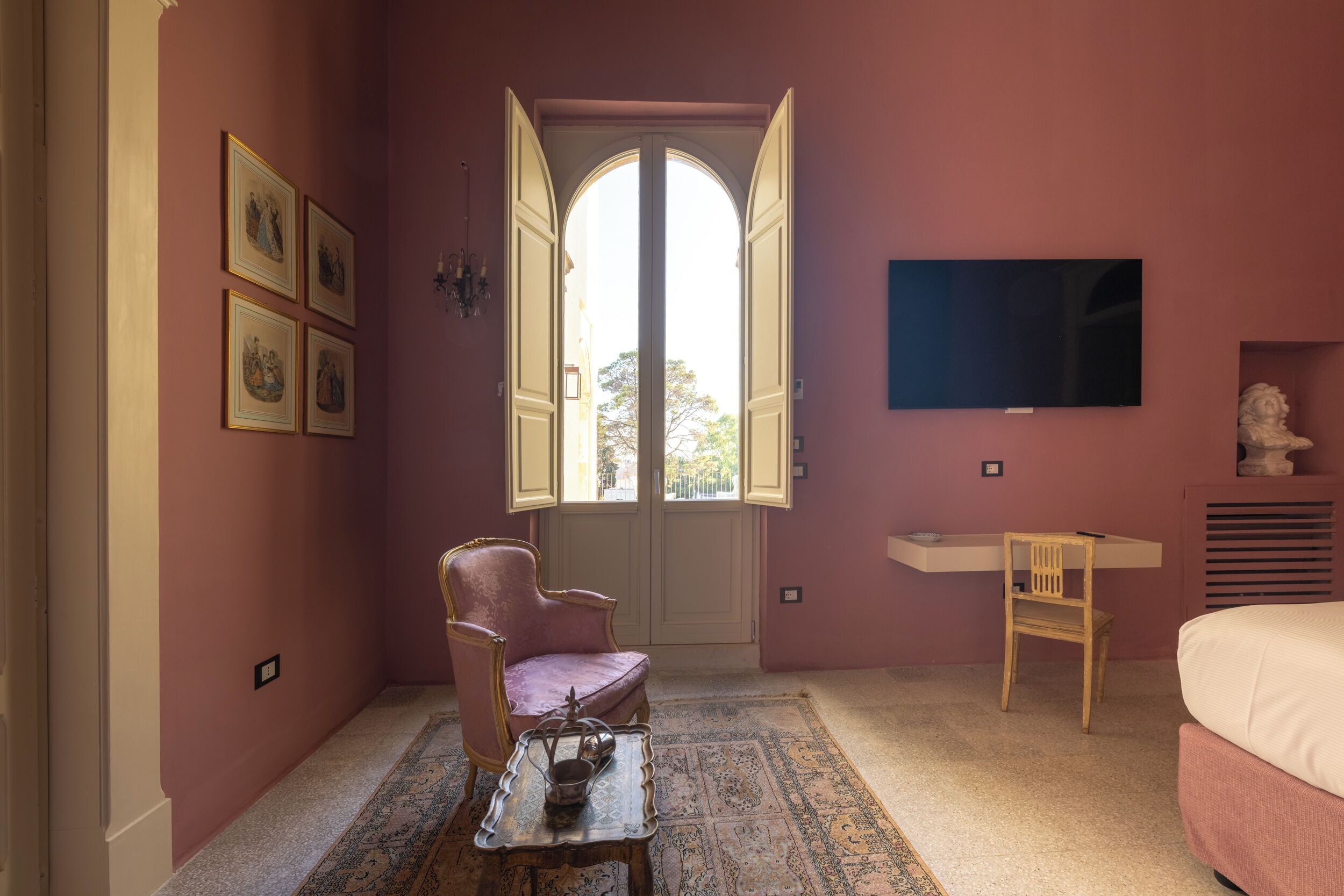 Una habitación con pared rosa, un ventanal arqueado grande, un televisor sobre un escritorio, una cama y una silla.