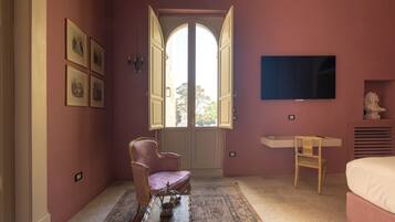 Una habitación con pared rosa, un ventanal arqueado grande, un televisor sobre un escritorio, una cama y una silla.