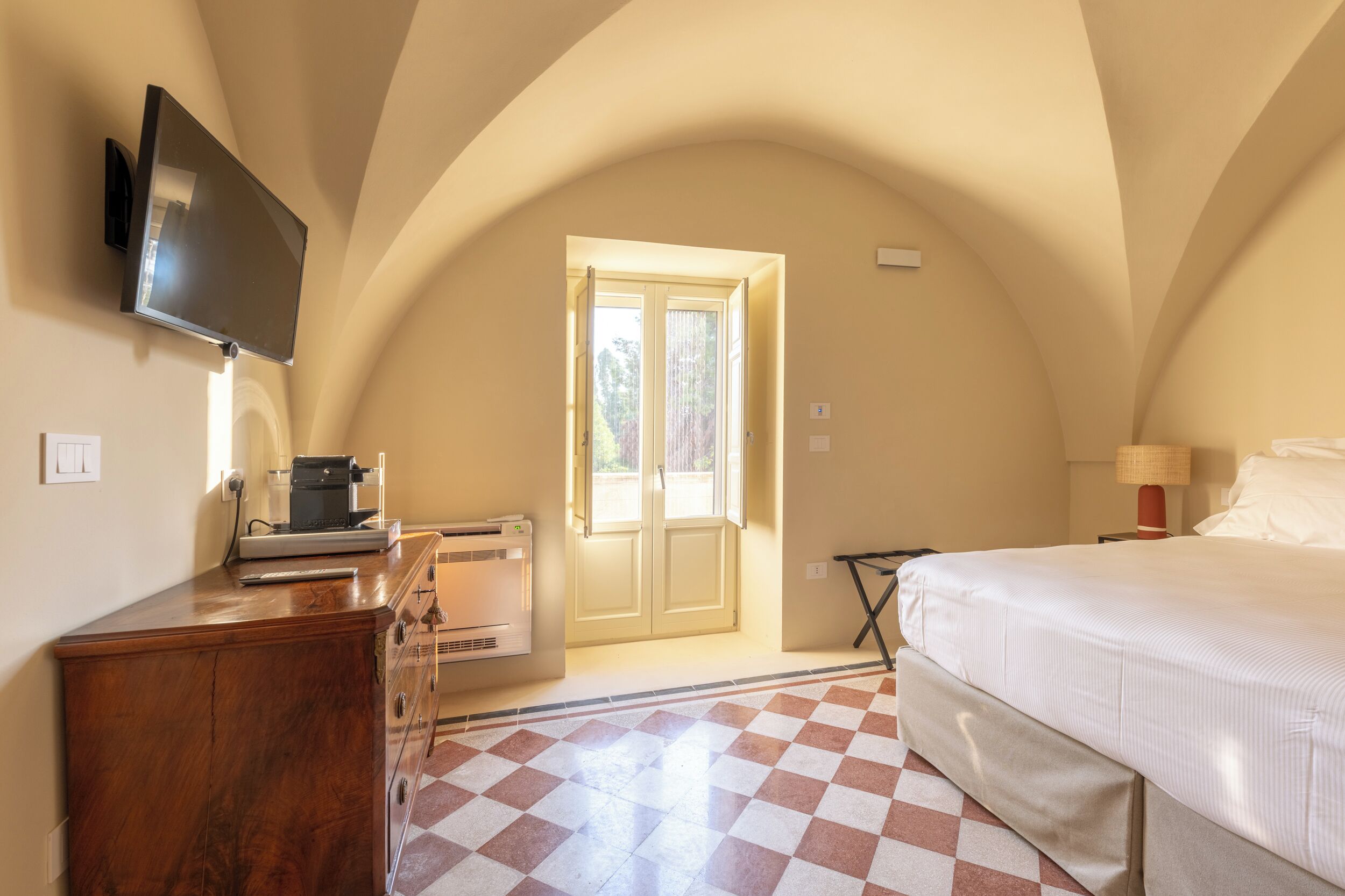 Una camera da letto con un letto, una TV a schermo piatto montata a parete, una scrivania con un computer e una finestra con tende.