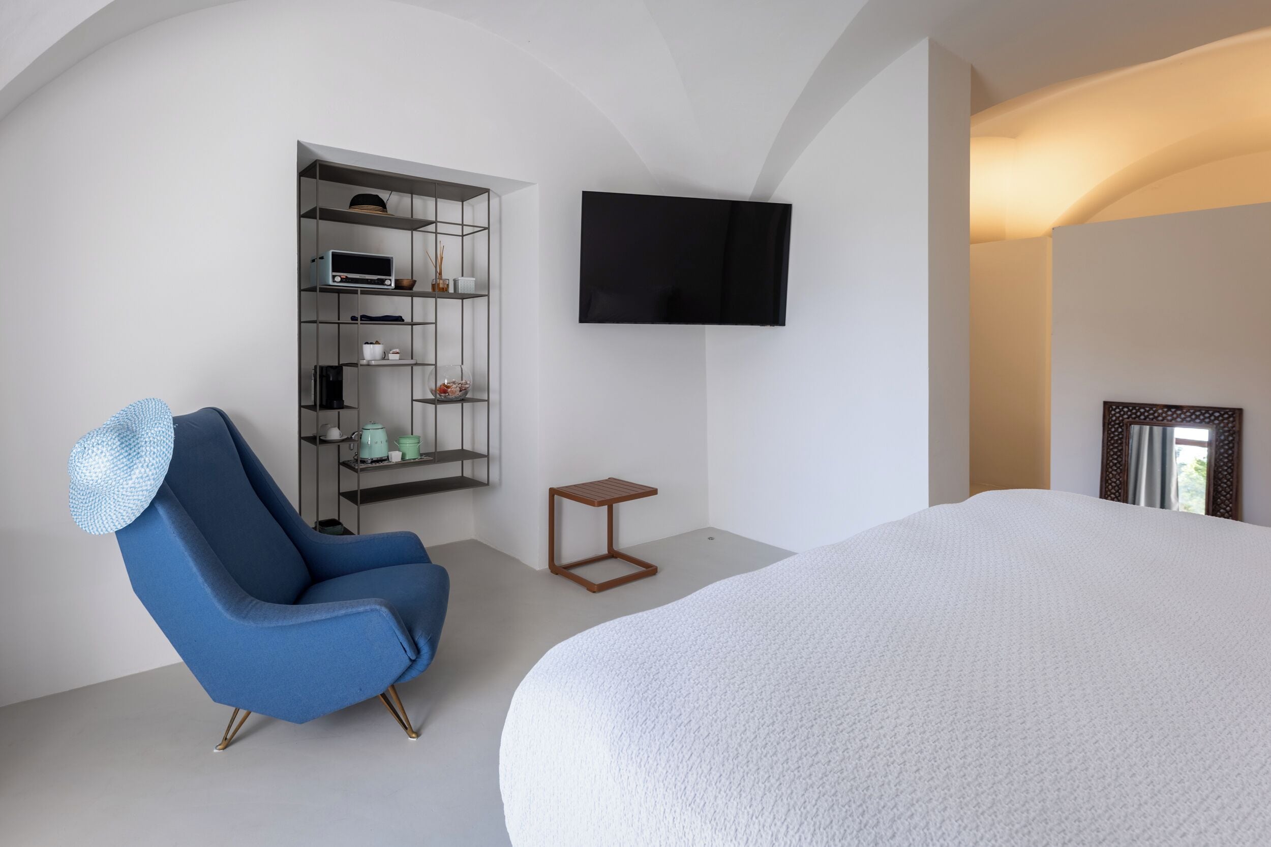 A modern bedroom with a large bed, a blue armchair, a TV mounted on the wall, and a built-in shelving unit.
