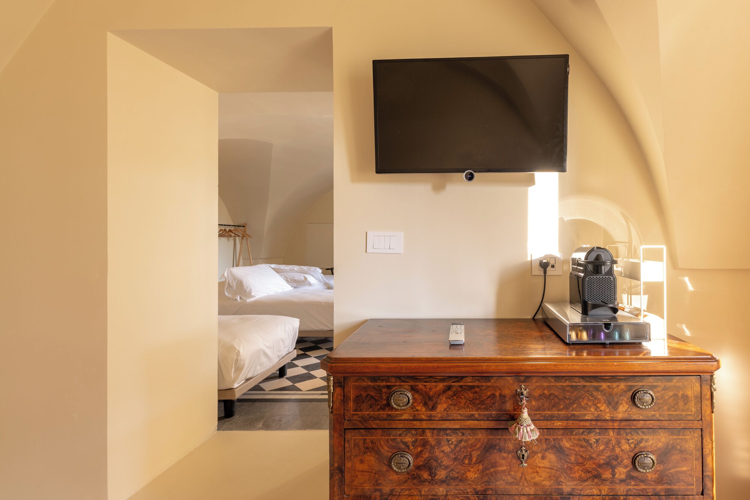 Une chambre d’hôtel avec une commode en bois, un téléviseur à écran plat fixé au mur, un lit avec une literie blanche et un sol à carreaux.