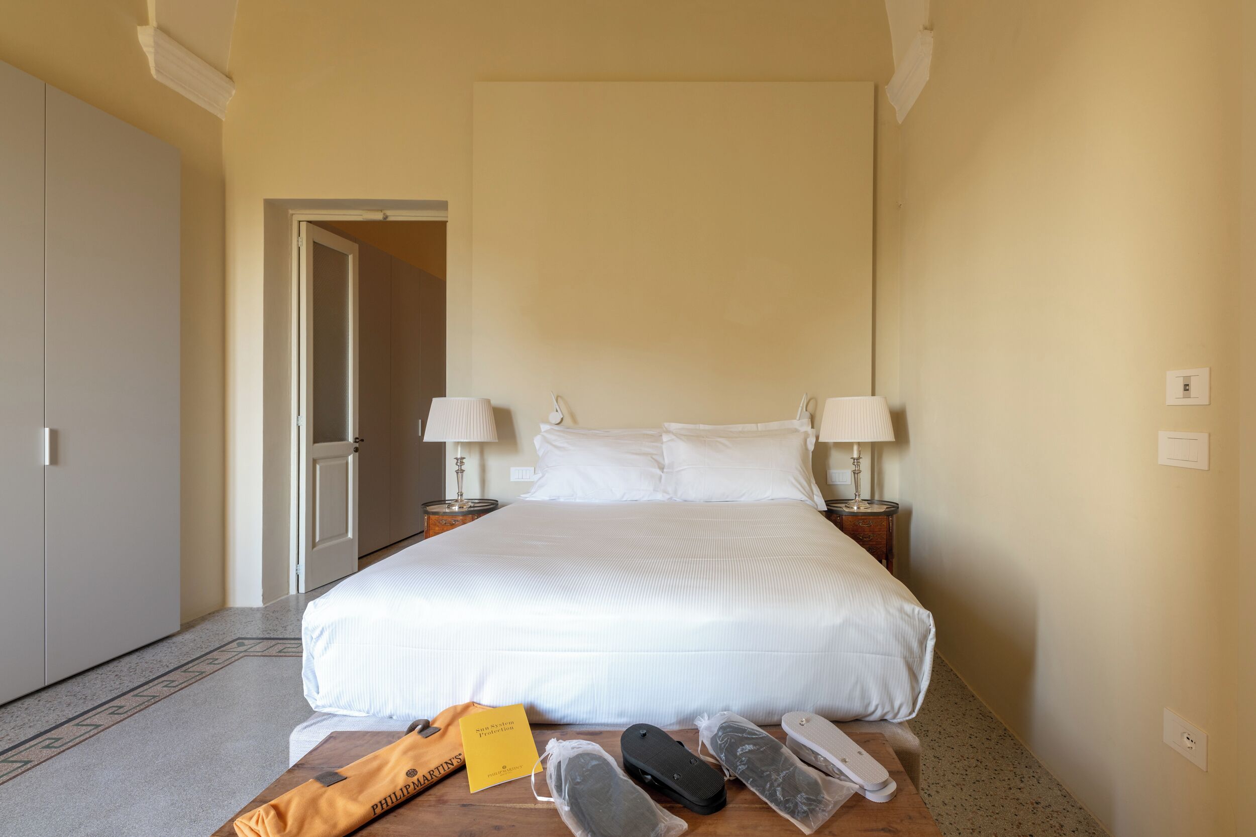 Ein Hotelzimmer mit einem großen Bett, zwei Nachttischen mit Lampen, einem Holzbeistelltisch und einer Tür zu einem weiteren Raum.