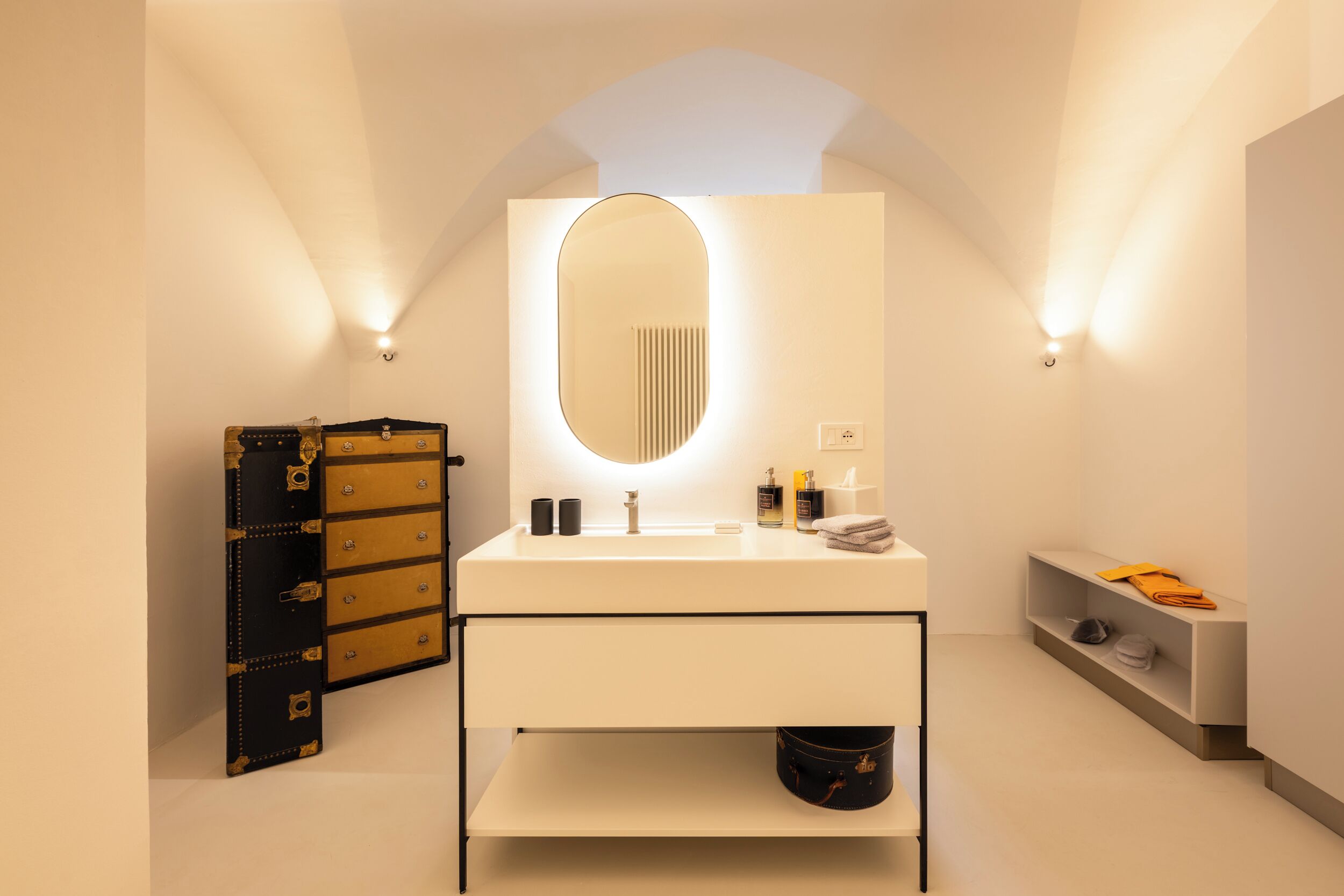 A modern bathroom with a white vanity, a large mirror, and a black and gold trunk.