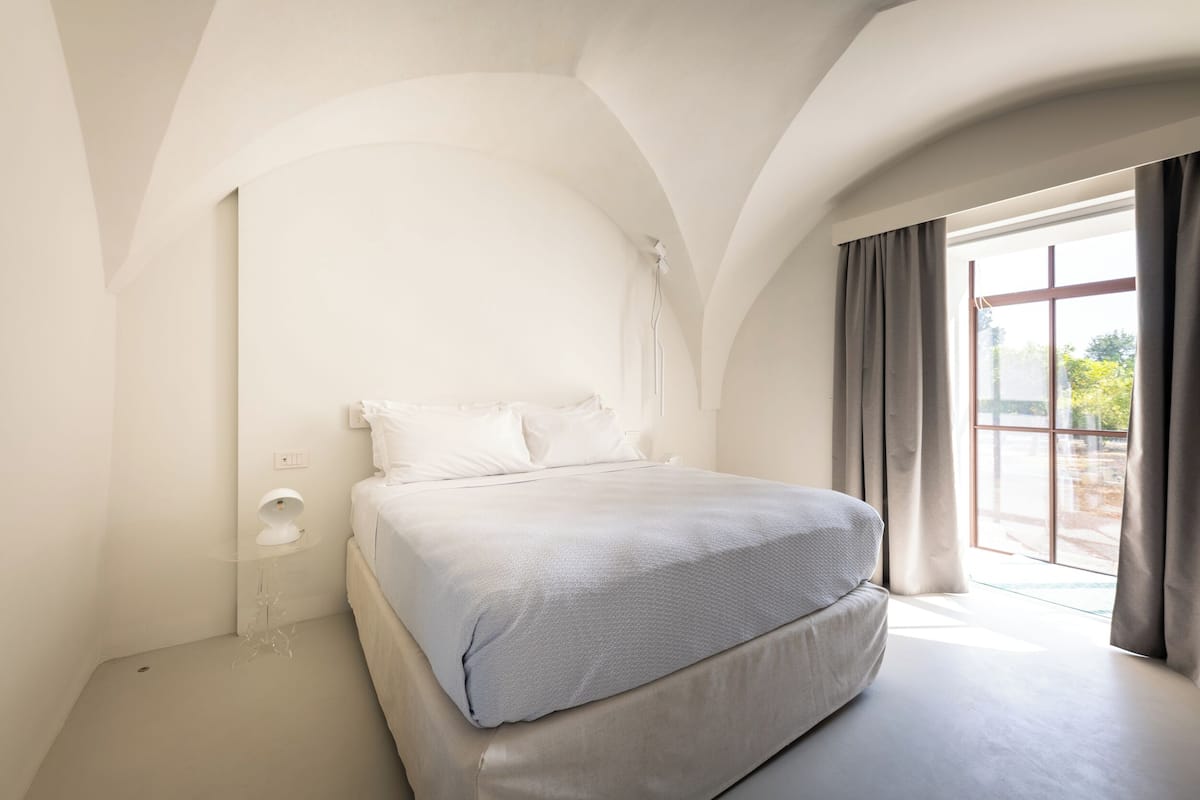 Un dormitorio con una cama grande, una mesita de noche con lámpara y una ventana con cortinas.