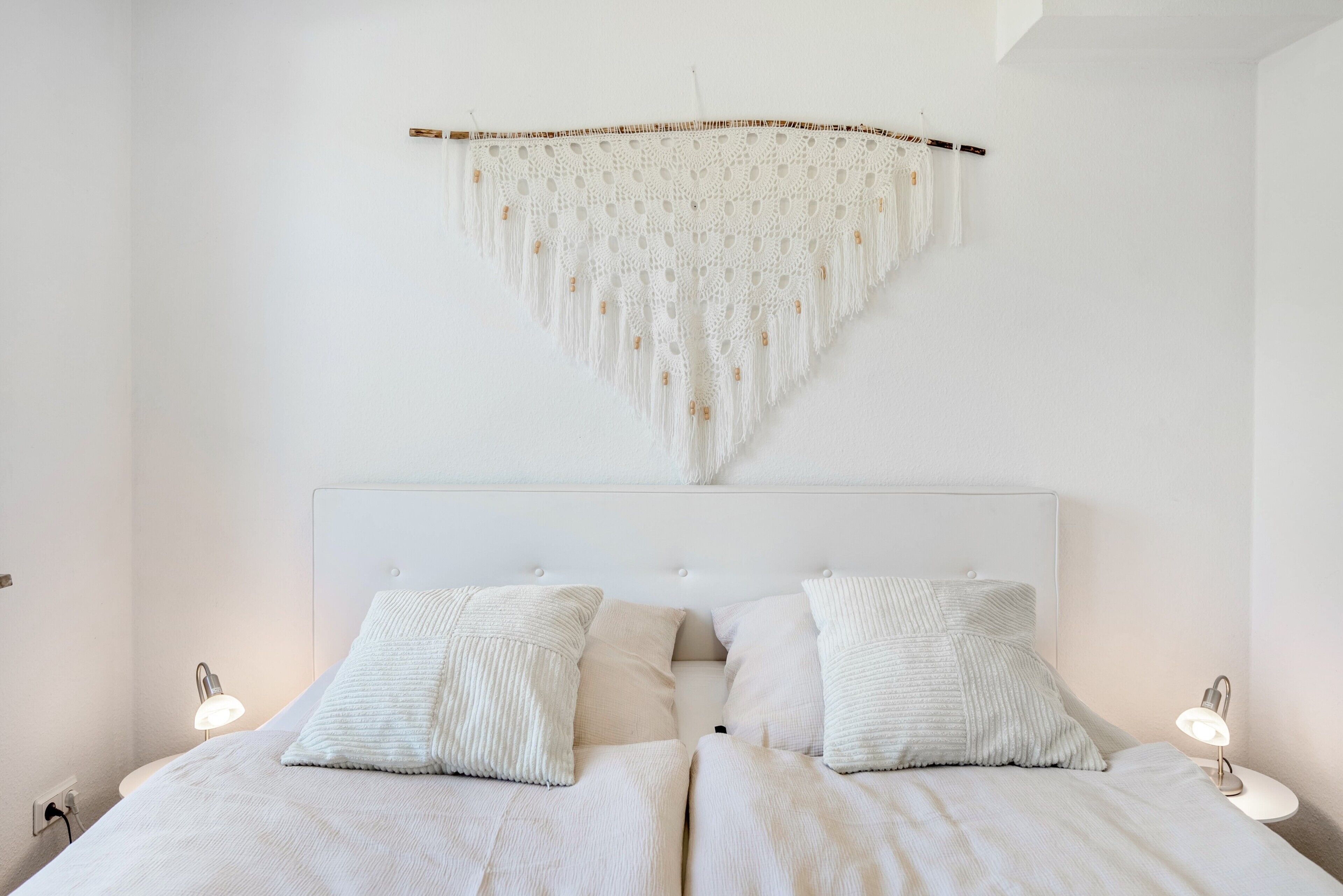 Een slaapkamer met een bed, twee bedlampen en een macramé wanddecoratie.