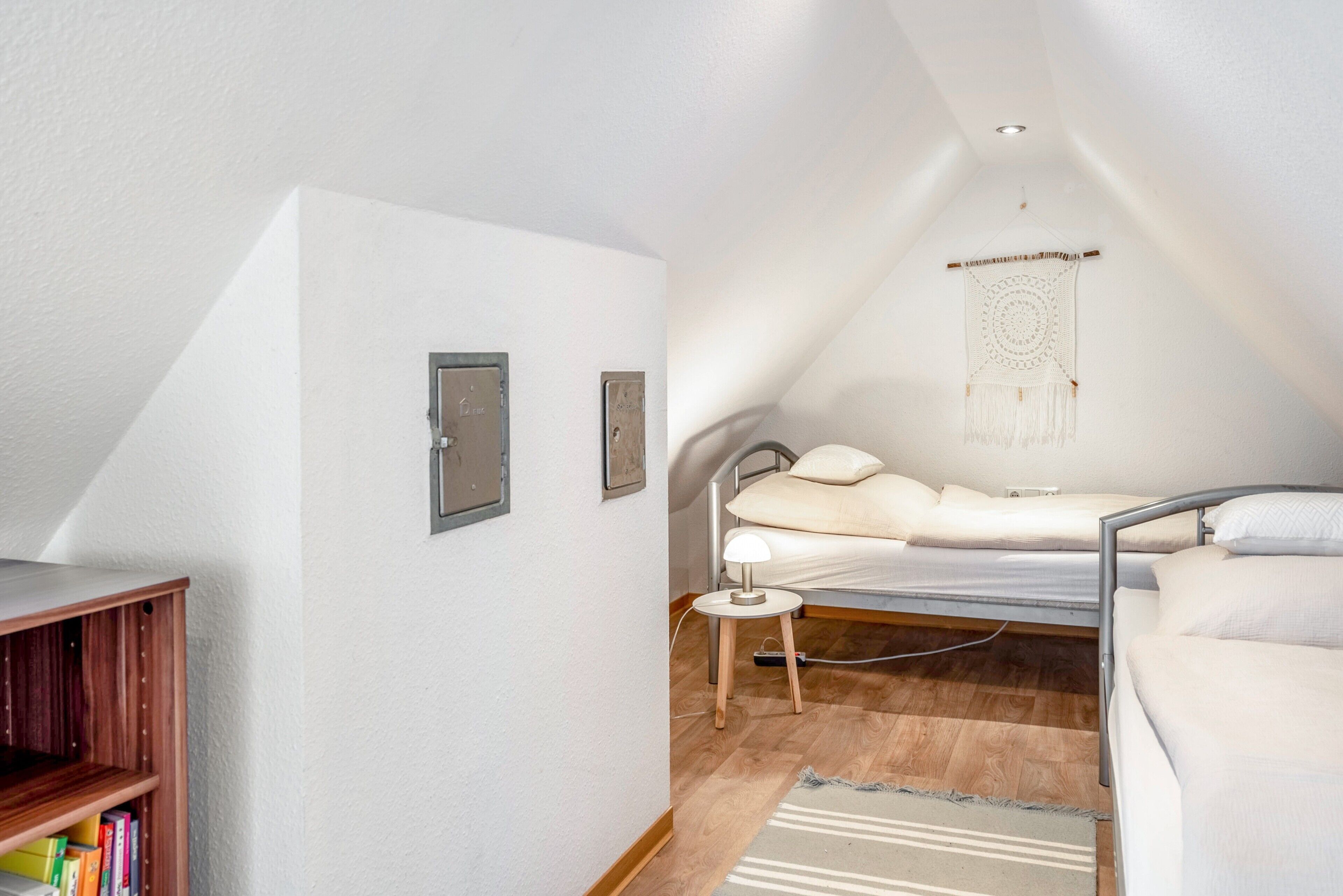 Una habitación pequeña y bien iluminada con techo inclinado, una cama con cabecera, una mesita de noche con lámpara, un tapiz colgado en la pared y una estantería.