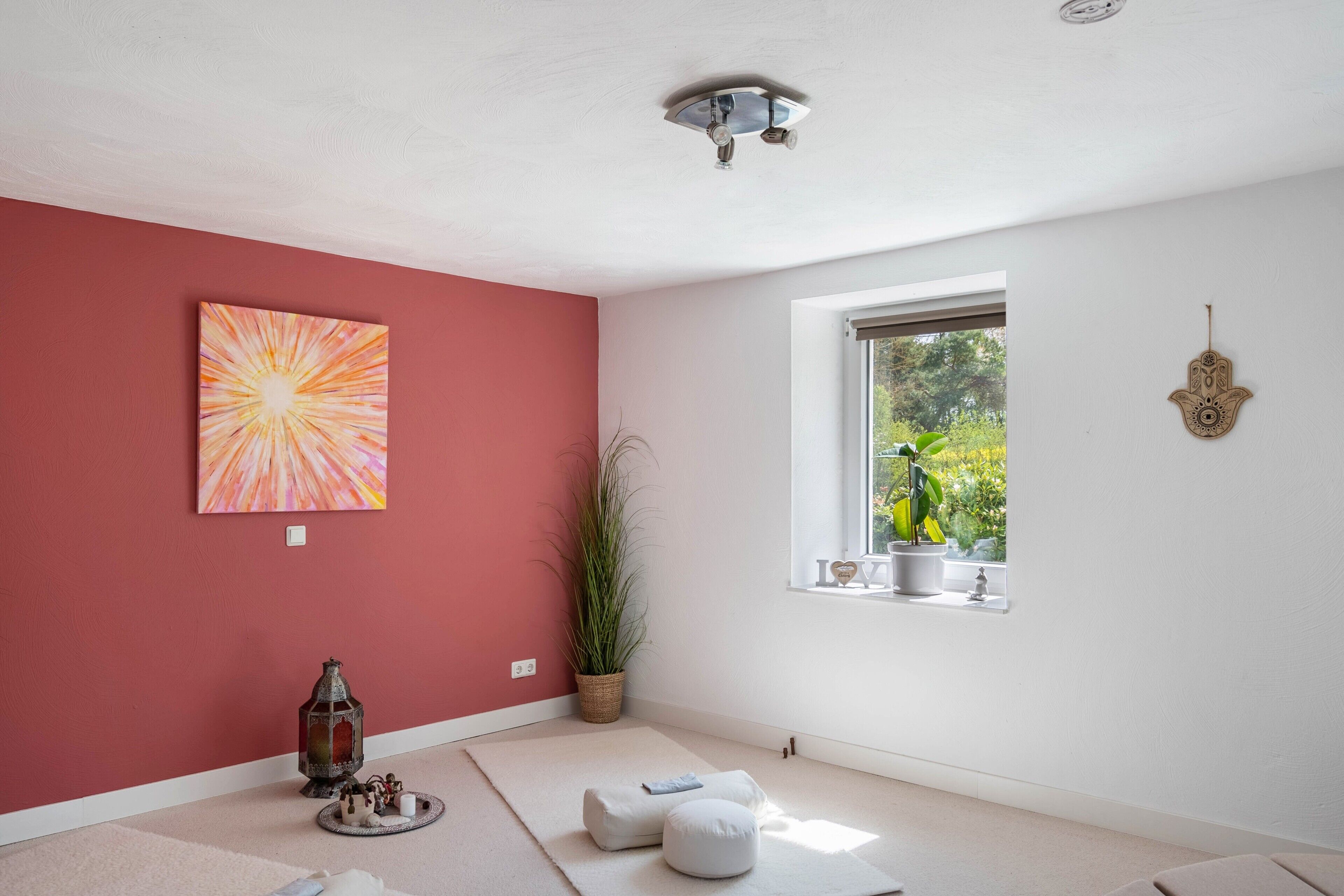 A room with a red wall, a large abstract painting, a window with a view of greenery, and a ceiling light fixture.
