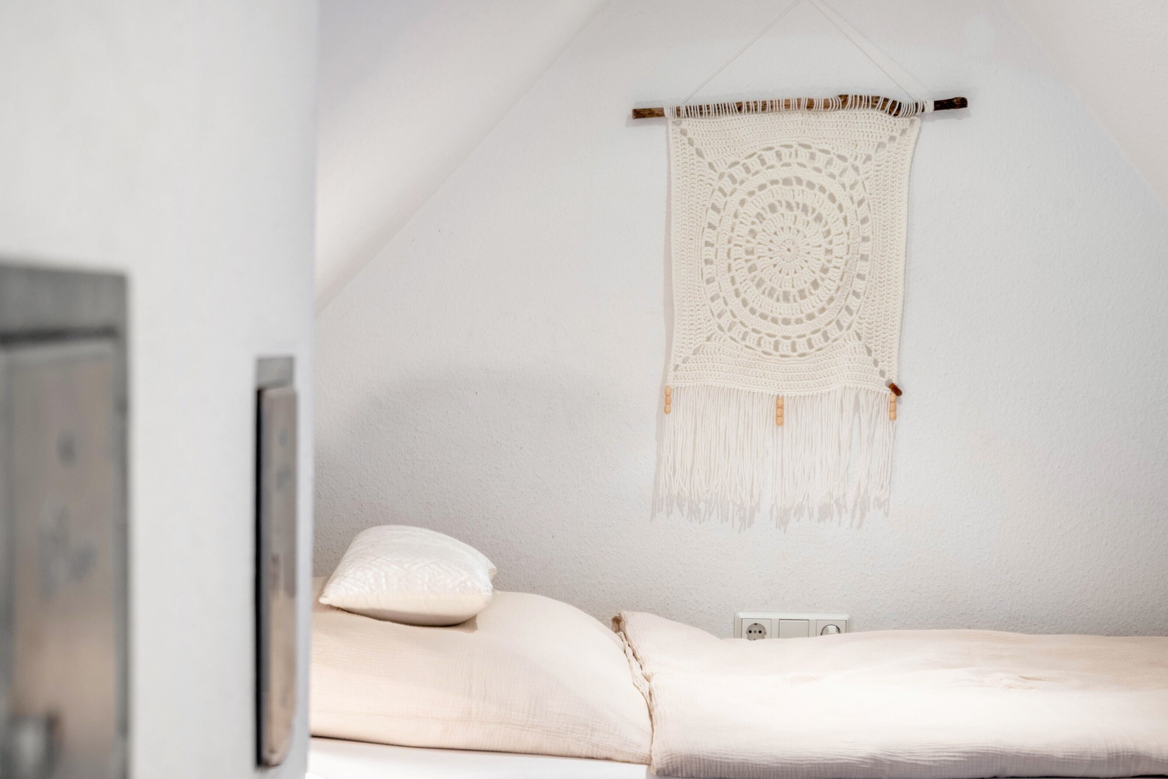 A bed with a white blanket, a pillow, and a wall-mounted macrame piece.