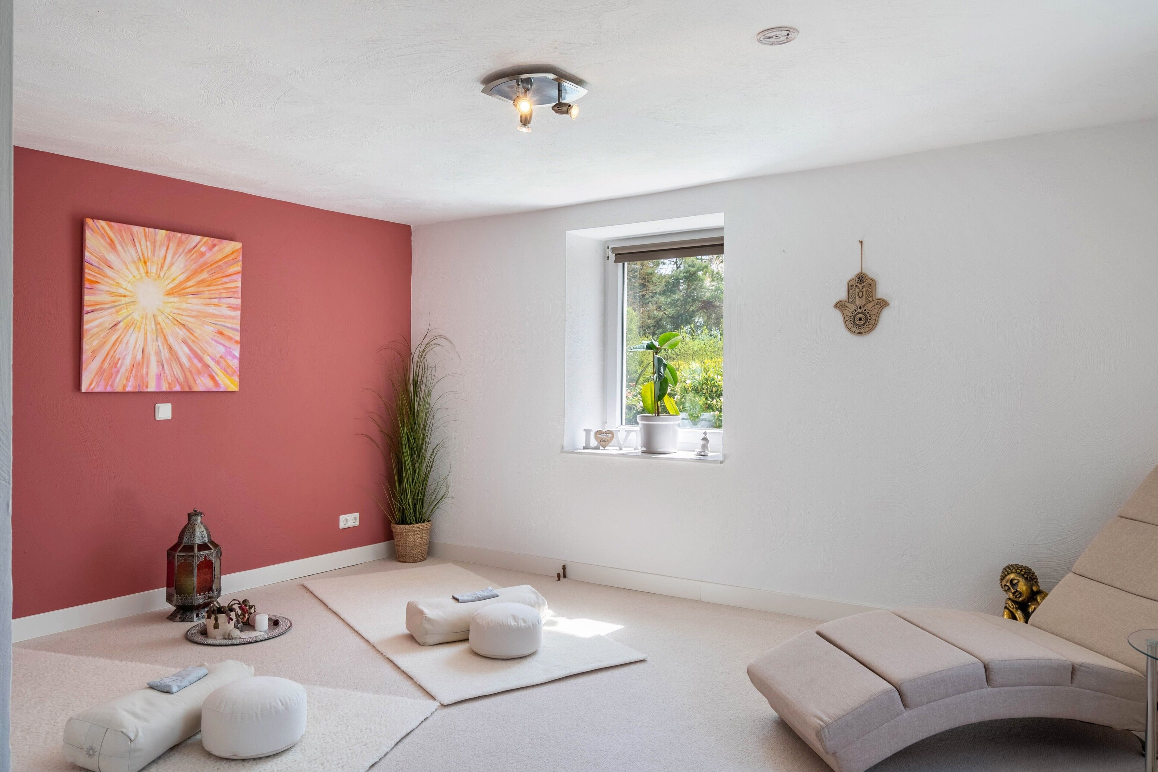A room with a red accent wall, a window with a view of greenery, a white sofa, and a floor lamp.