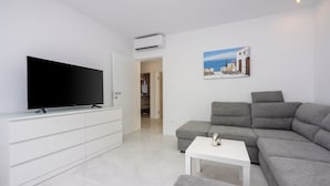 Ein modernes Wohnzimmer mit Flachbildfernseher, grauem Ecksofa, weißem Couchtisch und einem Gemälde einer Küstenlandschaft.