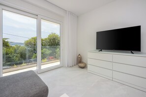 Ein modernes Zimmer mit großem Fenster, einem Flachbildfernseher, einem weißen Schrank und einem grauen Sofa.
