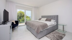 Ein modernes Schlafzimmer mit einem Bett, einem Glastische, einem Fernseher und Blick ins Freie.