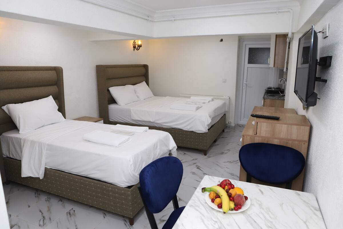 A hotel room with two beds, a desk with a chair, a small table with fruit, and a TV mounted on the wall.