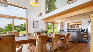 Un comedor amplio con una mesa y sillas de madera, ventanas grandes y una cocina al fondo.