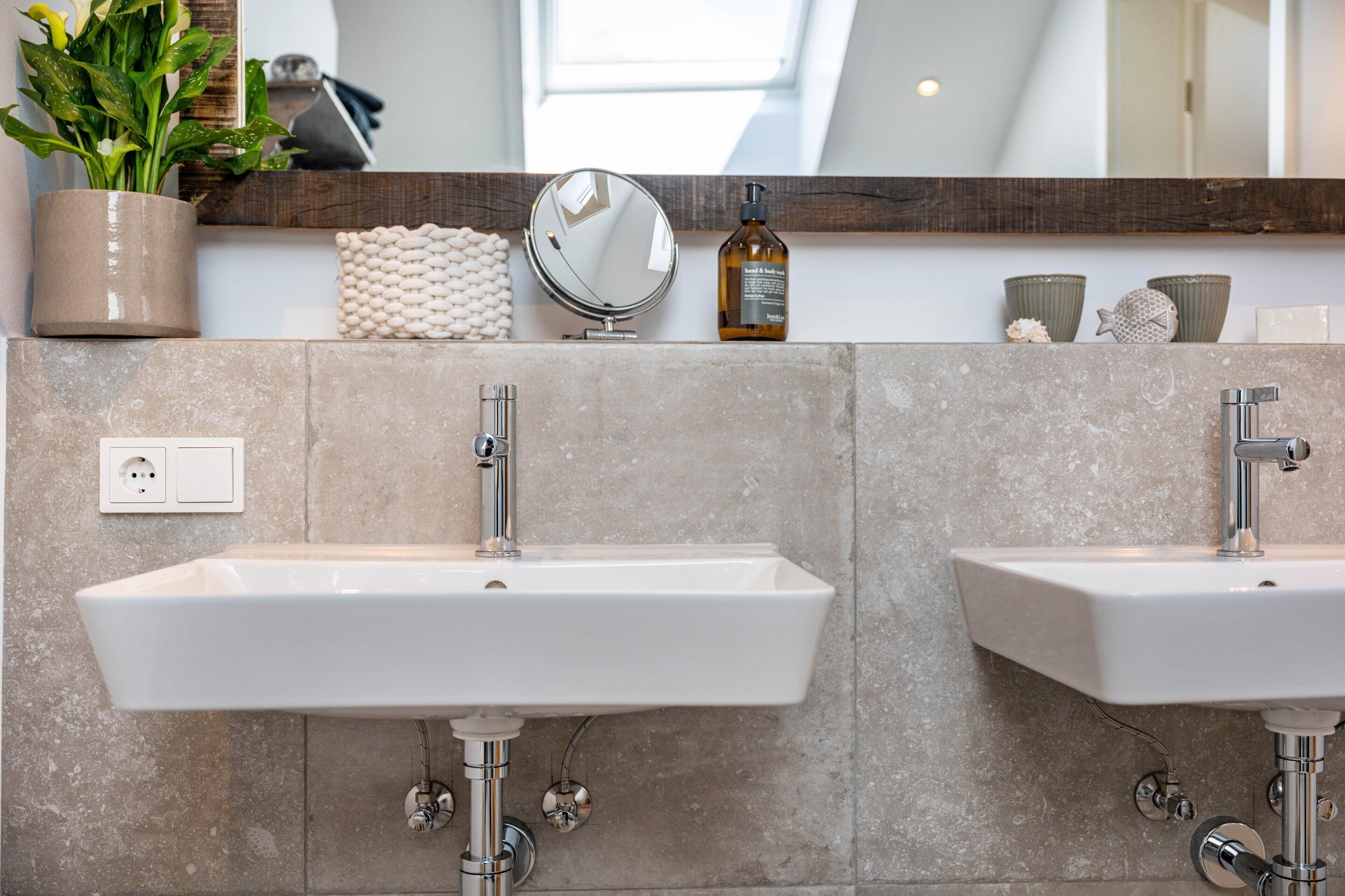 A bathroom with two sinks, a mirror, a potted plant, and a shelf with toiletries.