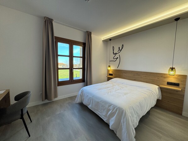 A modern bedroom with a bed, a desk, a chair, a window with curtains, and a wall with a light fixture.