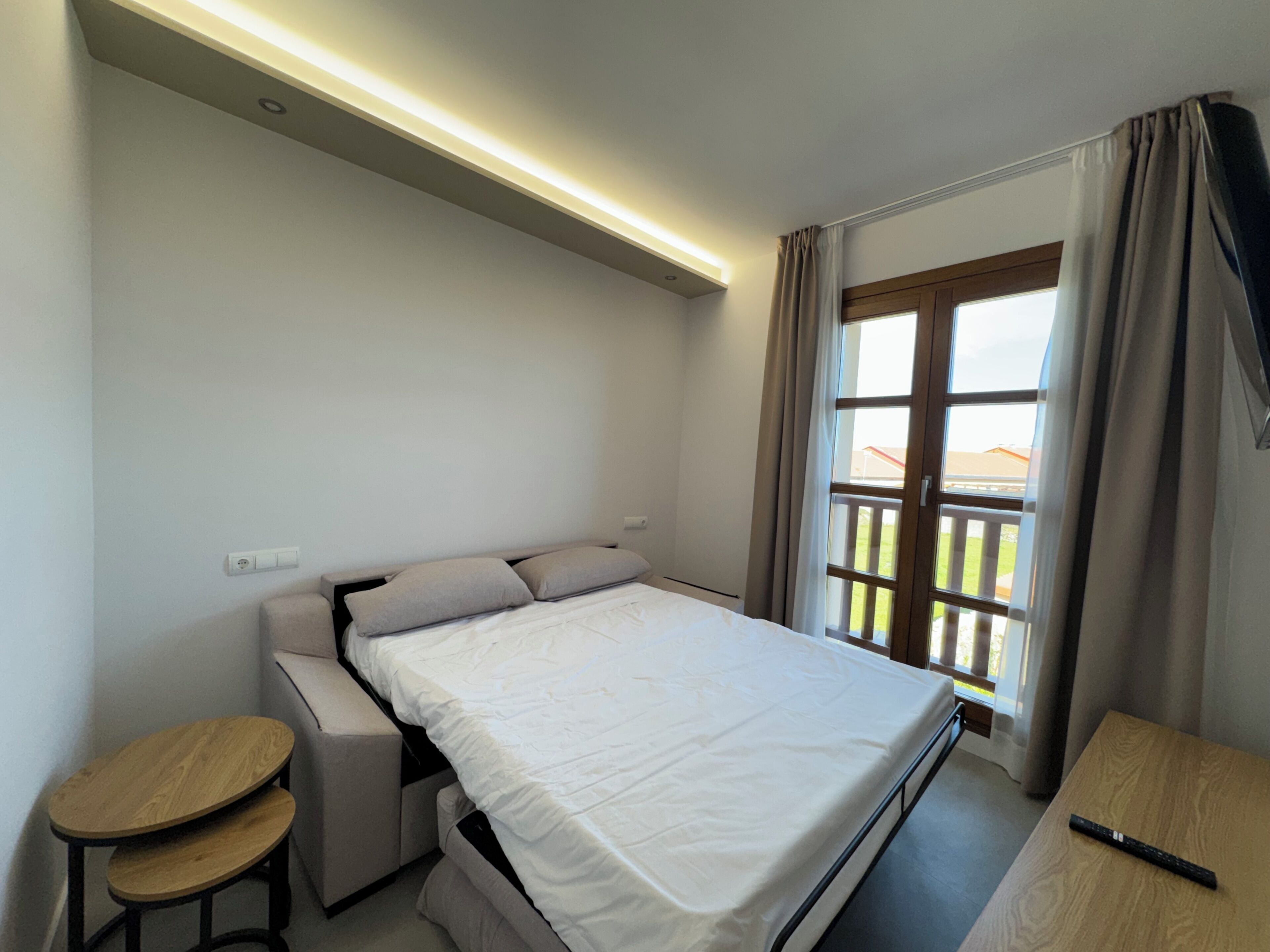 Un dormitorio con cama, mesitas de noche, escritorio, ventana con cortinas e iluminación en el techo.