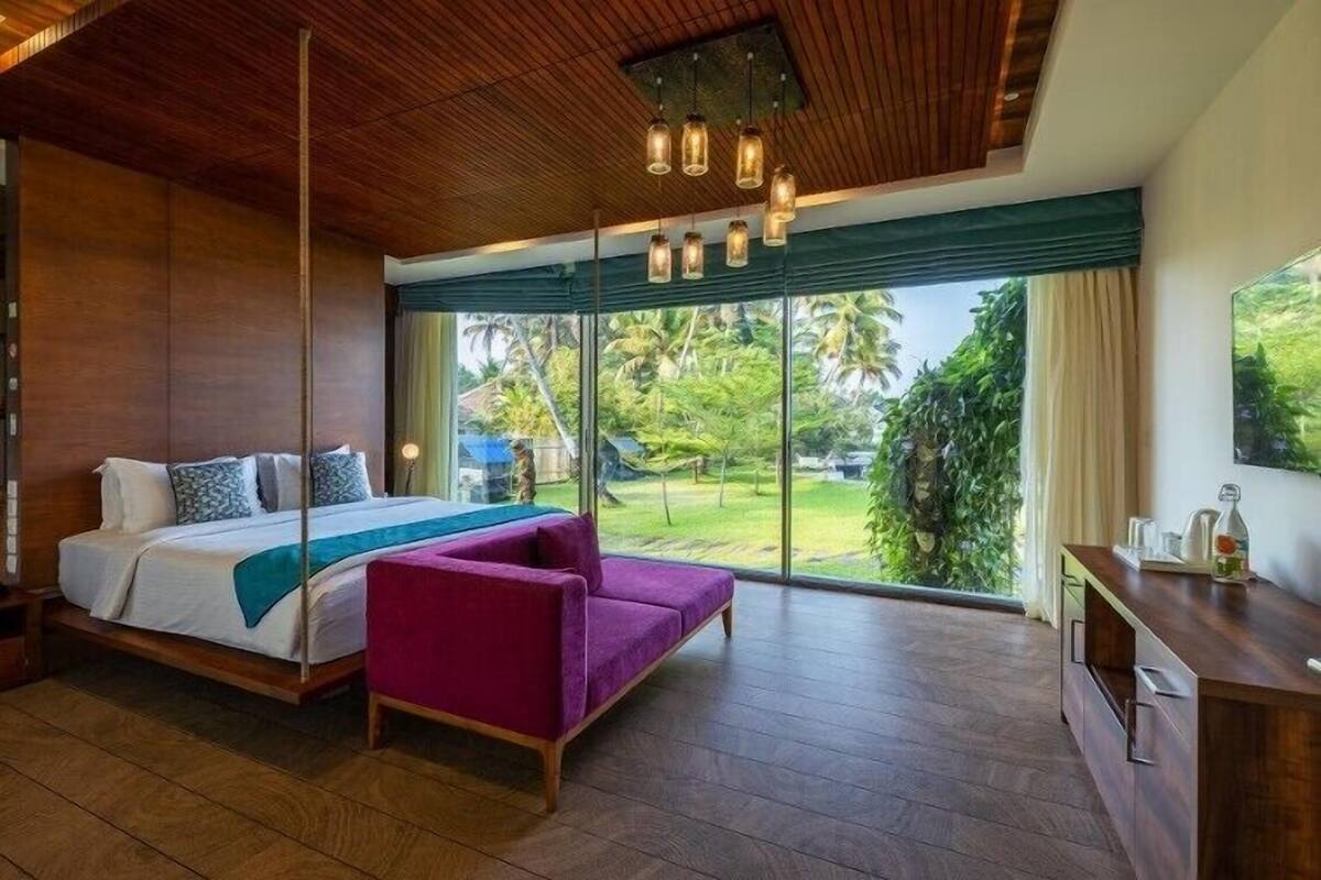 Ein modernes Hotelzimmer mit einem großen Bett, einem violetten Sessel, einer Holzbalkendecke und Blick ins Grüne.