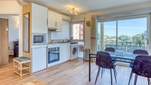 A compact kitchen with a microwave, oven, and washing machine, a dining area with a glass table and chairs, and a large window with a view of buildings and greenery. - Cosy Renovated Apartment Near the Beach With Balcony (Lattes)