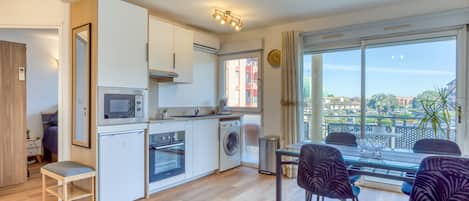 A compact kitchen with a microwave, oven, and washing machine, a dining area with a glass table and chairs, and a large window with a view of buildings and greenery.