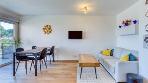 A modern living room with a dining area, a sofa, a TV mounted on the wall, and a large window. - Cosy Renovated Apartment Near the Beach With Balcony (Lattes)