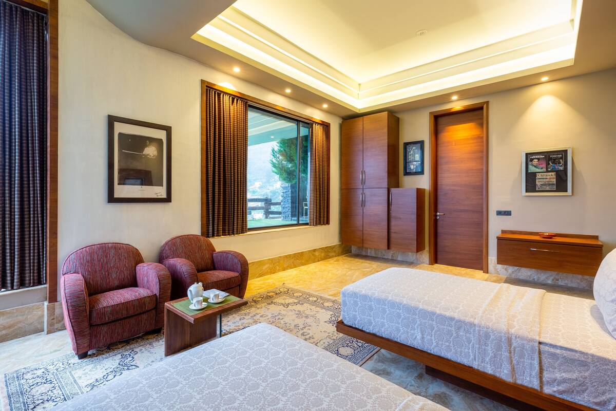 Ein Schlafzimmer mit einem Bett, zwei Sesseln, einem kleinen Tisch, einem Kleiderschrank und einem Fenster mit Vorhängen.