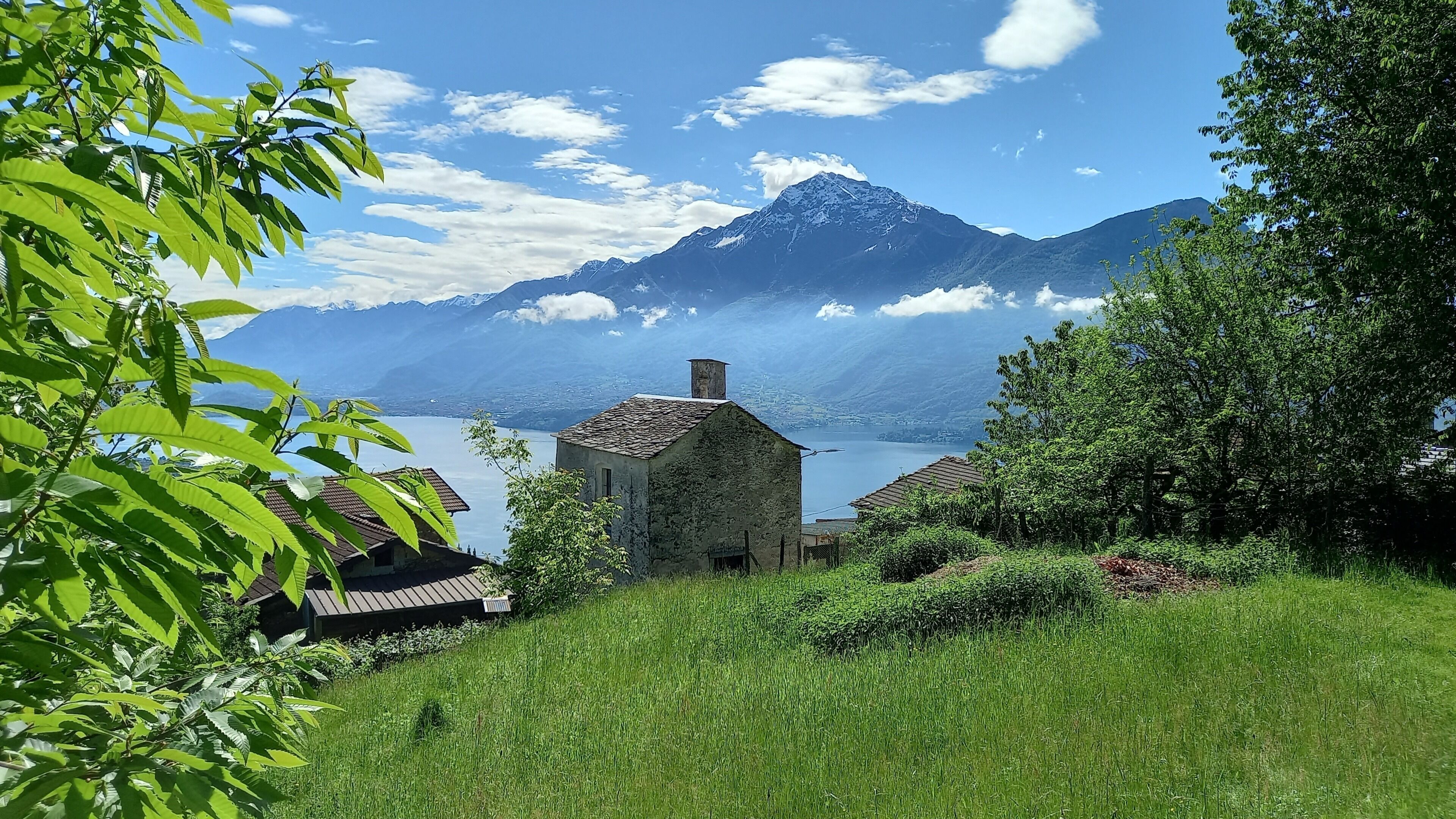 A stone building with a sloped roof, surrounded by lush greenery and a lake, with mountains in the background.